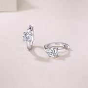A pair of Éclat Solitaire Moissanite Huggie Earrings made with sterling silver and featuring round-cut moissanite stones, displayed against a neutral background.
